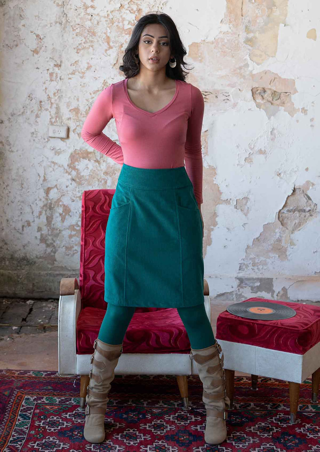 Model wears a knee length A-line corduroy skirt with wide waistband and pockets, paired with a pink top and teal leggings and boots