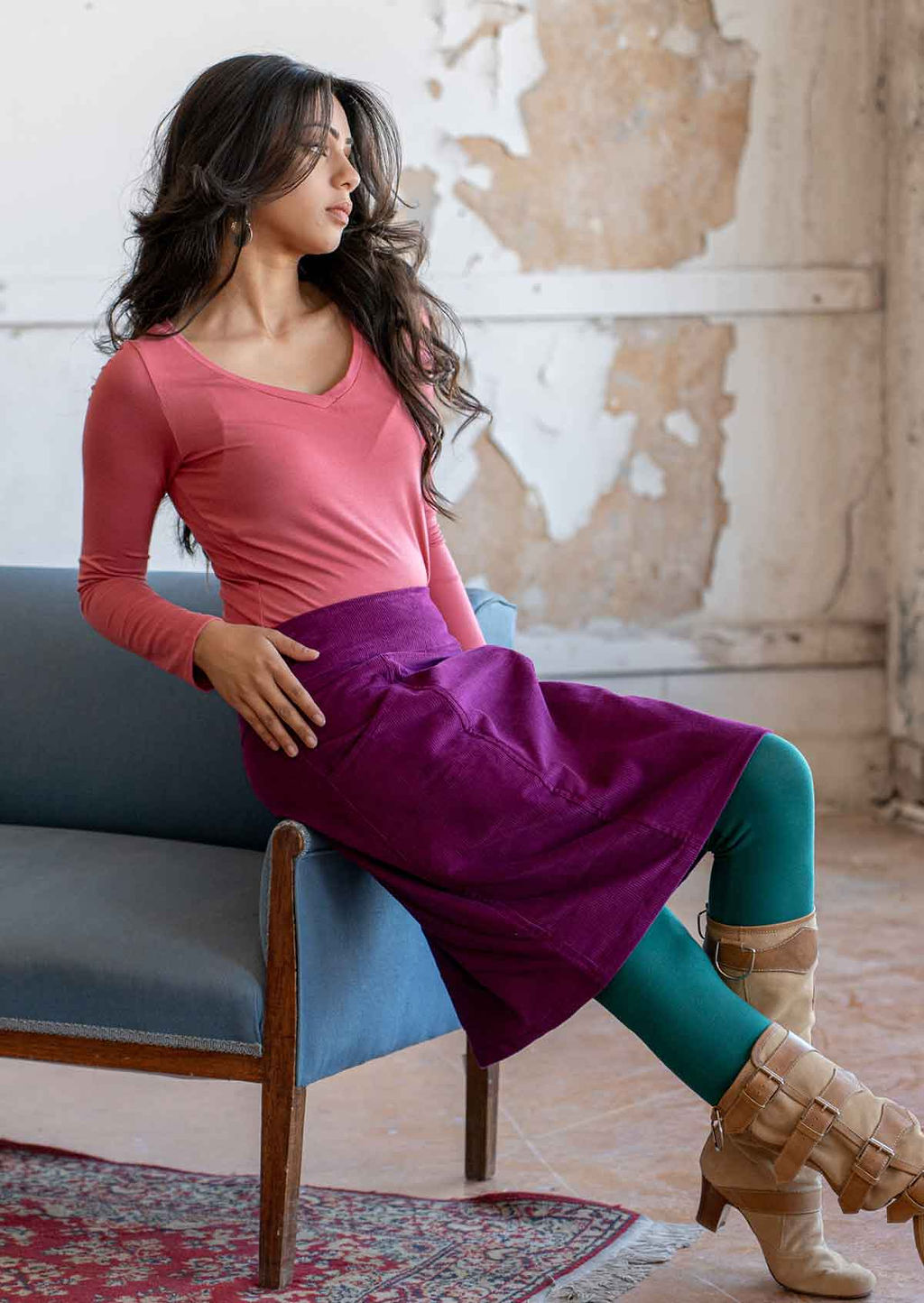 Woman reclines on arm of chair in a purple corduroy skirt paired with a long sleeve pink top and teal leggings and boots
