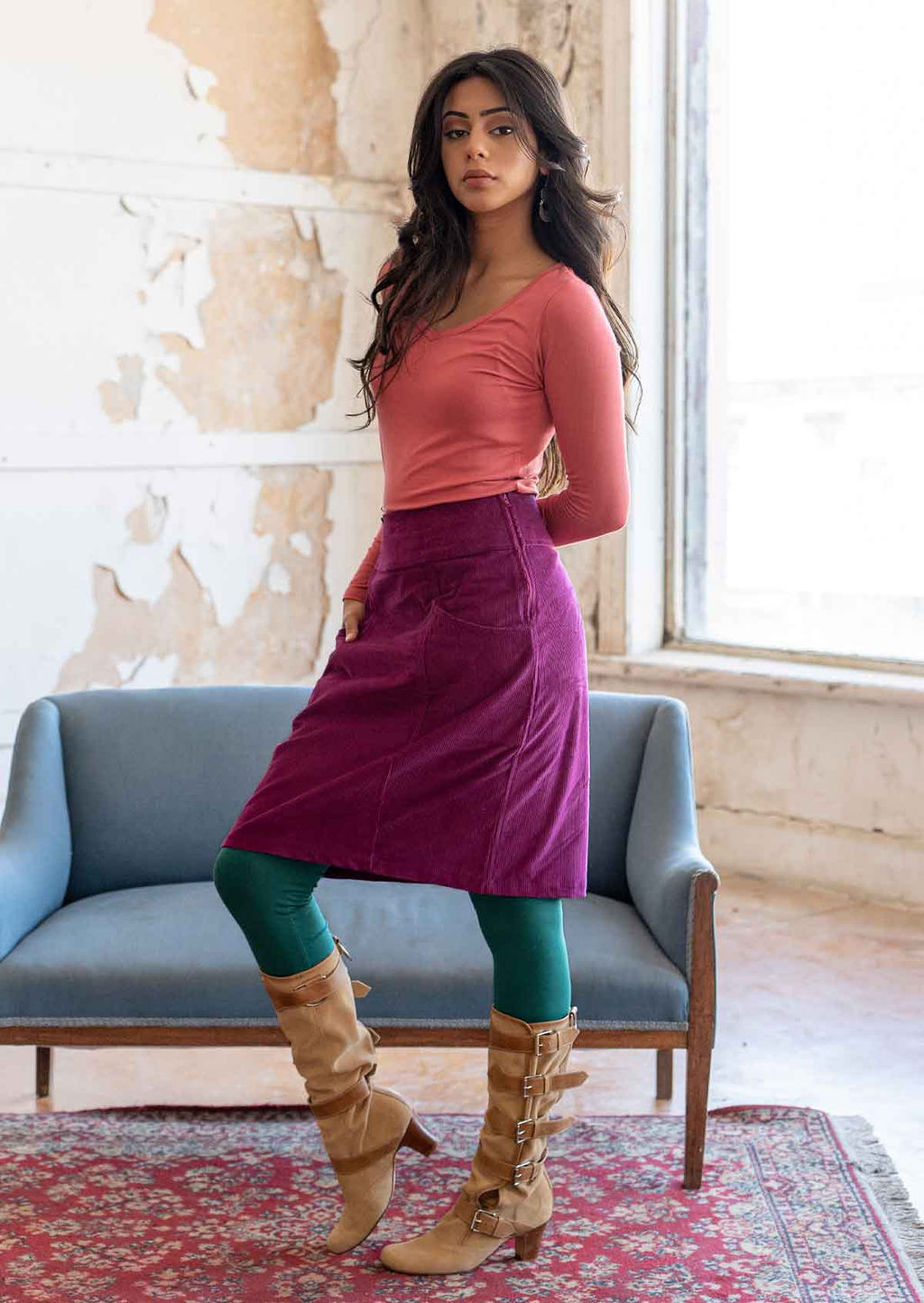 Woman stands in corduroy A-line knee length skirt with a hand in her pocket, paired with a pink long sleeve top