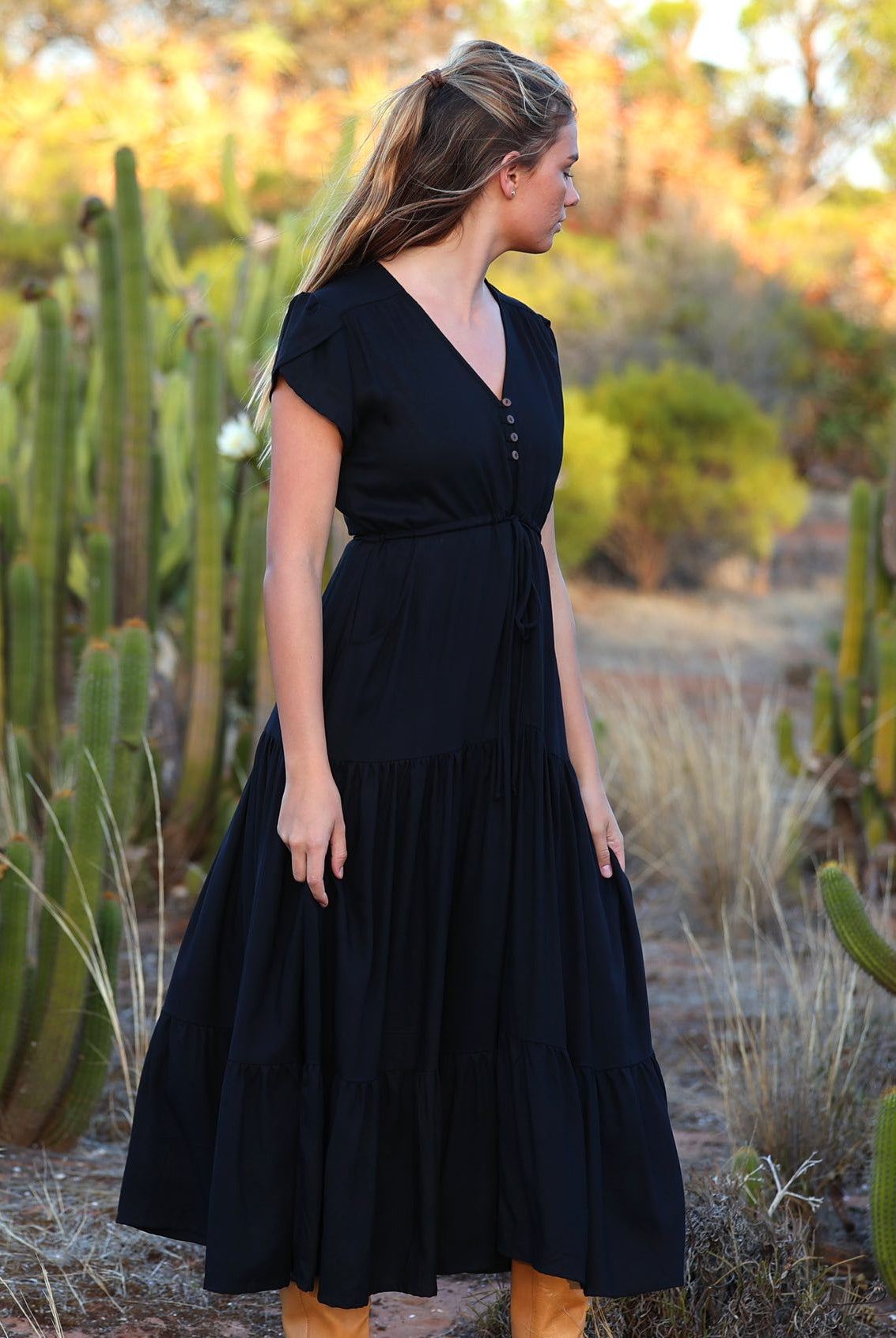 Woman stands in black rayon maxi dress with tulip sleeves, V-neckline and buttoned bodice