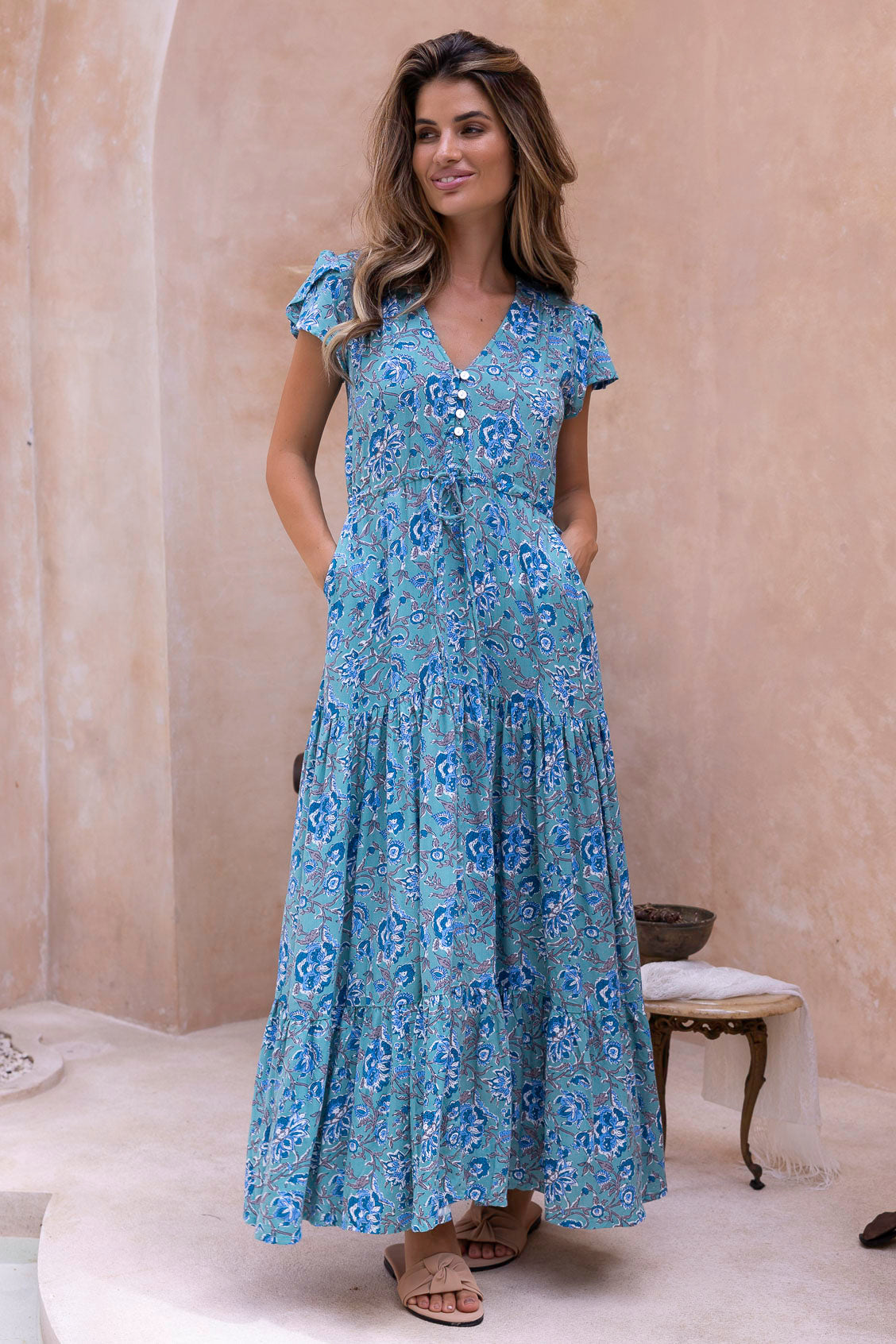 Woman wearing a beautiful blue floral dress perfect for wedding guest standing in a room with beige walls and a white rug.