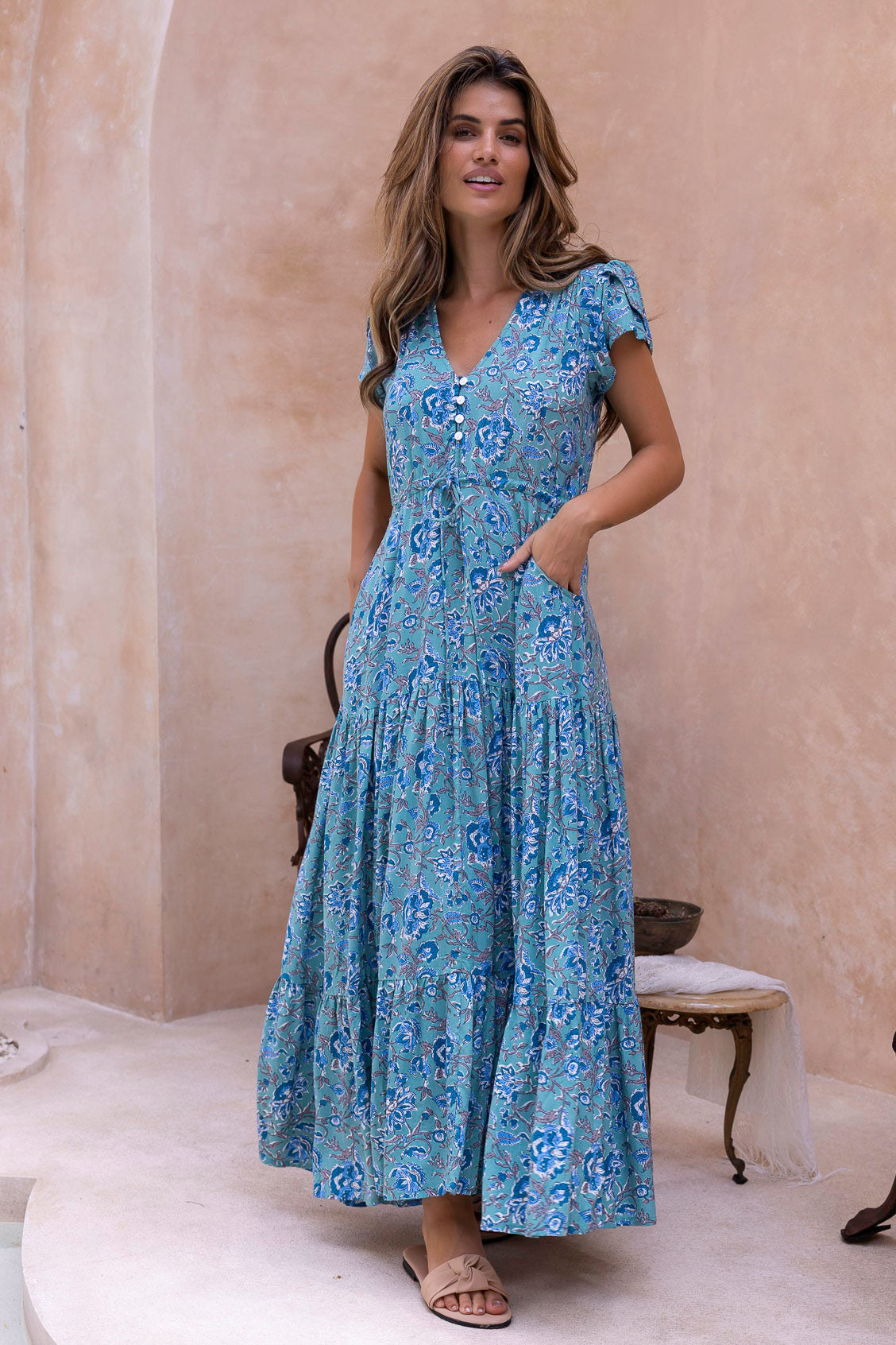 Woman wearing a blue floral maxi dress standing against a beige wall with hand in pocket