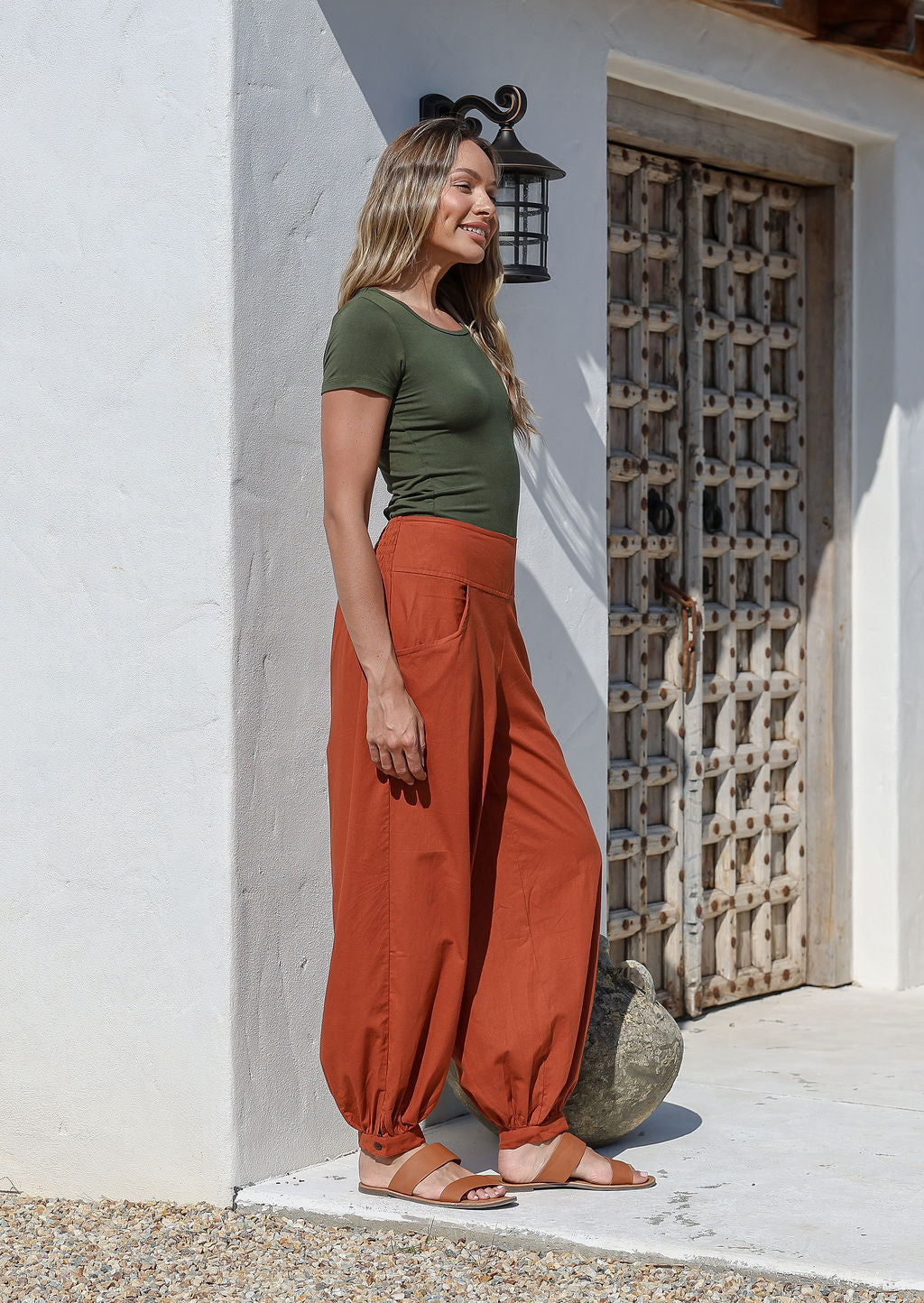 A woman wearing a green basic top and brick orange, genie style pull-on cotton pants with pockets, cuffed buttoned ankles, and a shirred elastic waistband.
