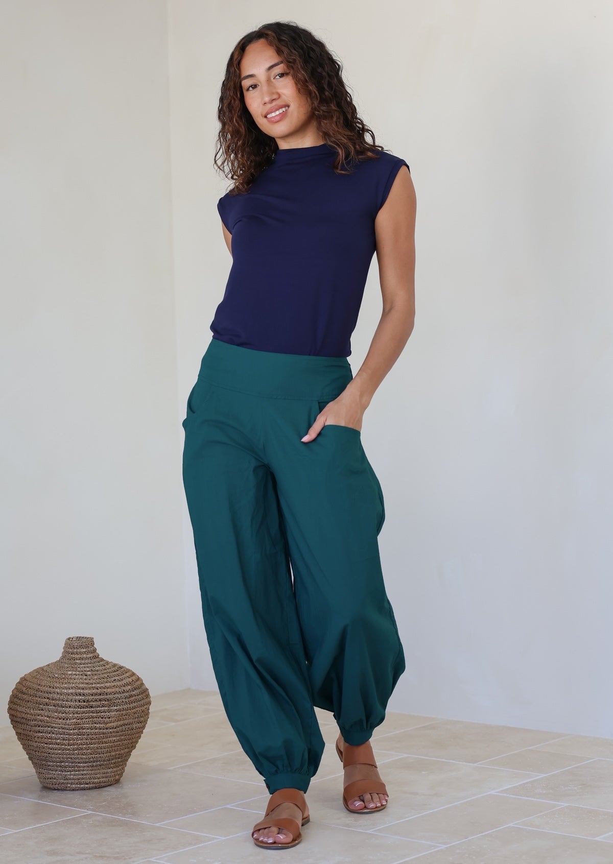 Woman stands with one hand in pocket wearing a genie style pull on pants in deep teal with cuffed ankles, wide leg fit and flat wide waist band that is shirred at the back