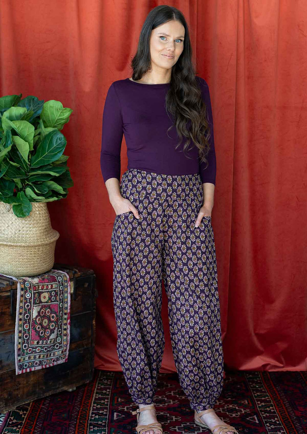 Woman wears cotton buttoned ankle harem pants with floral motif print on a dark purple base