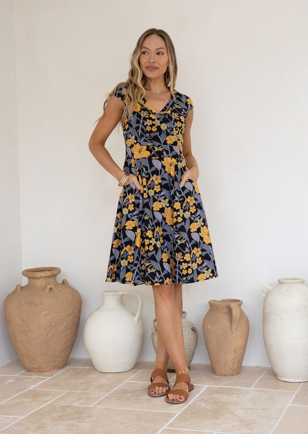 Model with both the hands in pocket wears 100% cotton dress with crossover bodice, cap sleeves and fuller skirt in retro  floral print.