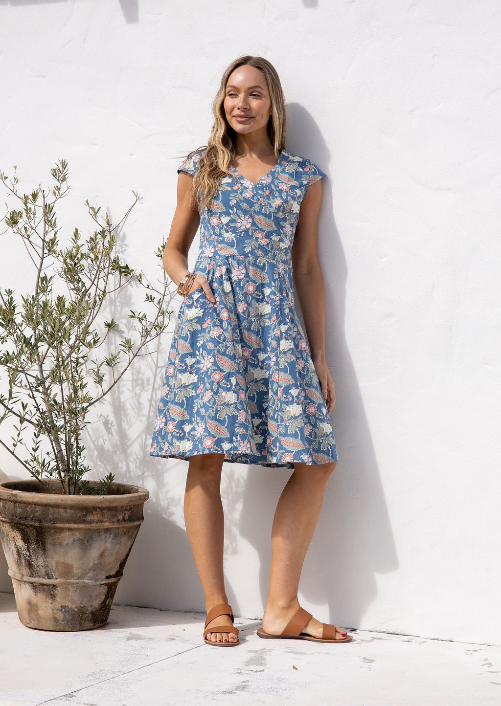 Model standing infront of white wall wears above knee length cotton dress with crossover bodice, cap sleeves and A line skirt.