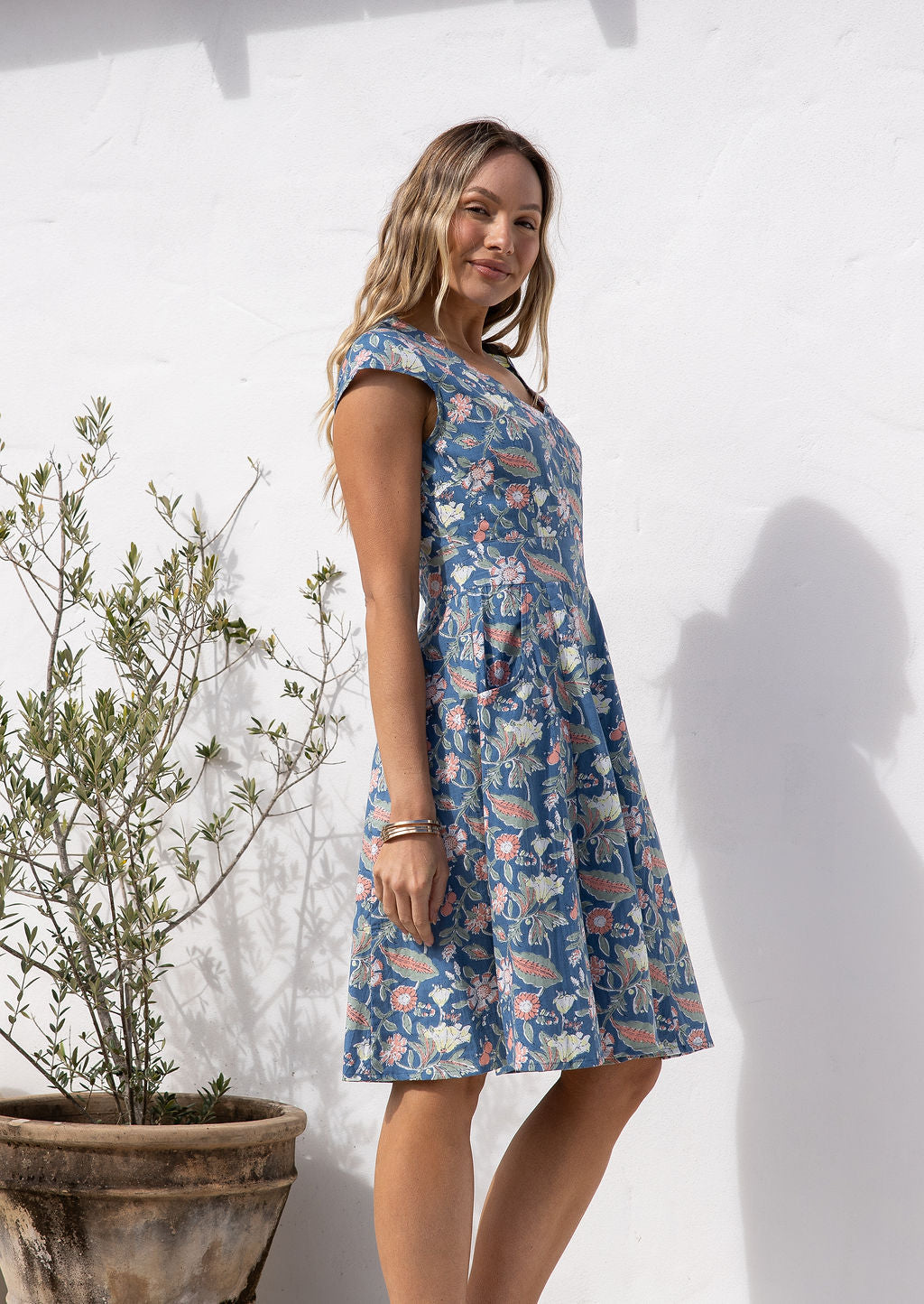 Woman stands in sun wearing a gorgeous floral cotton dress in dusty blue with fit and flare style and pockets.