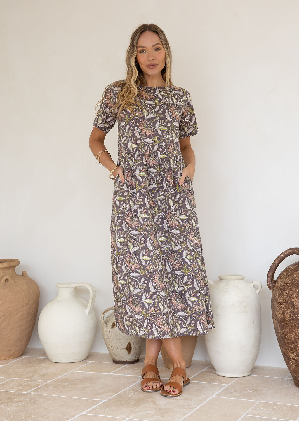 Woman standing in front of decorative vases wears 100 percent cotton relaxed fit, midlength dress in taupe based coral pink, lime and beige froral print.