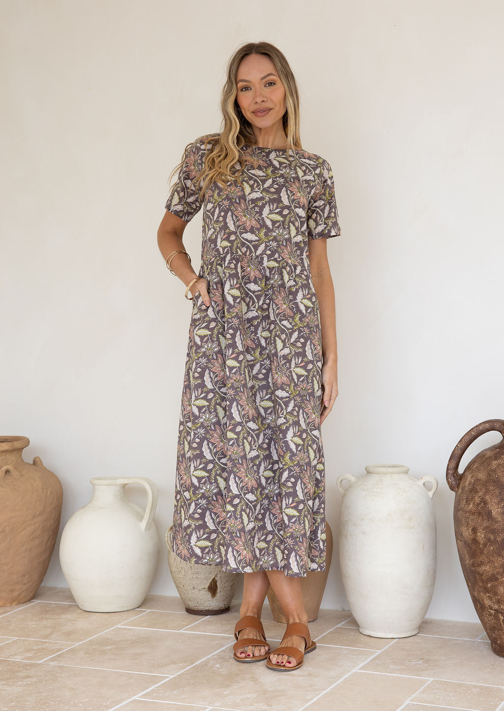 Woman poses with one hand in pocket of short sleeved midlength cotton dress with boat neckline and taupe based delicate floral print.