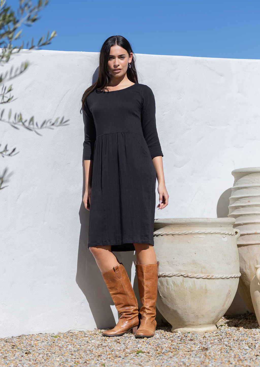 Woman wearing a black 100 cotton dress and brown boots standing against a white wall with decorative urns.