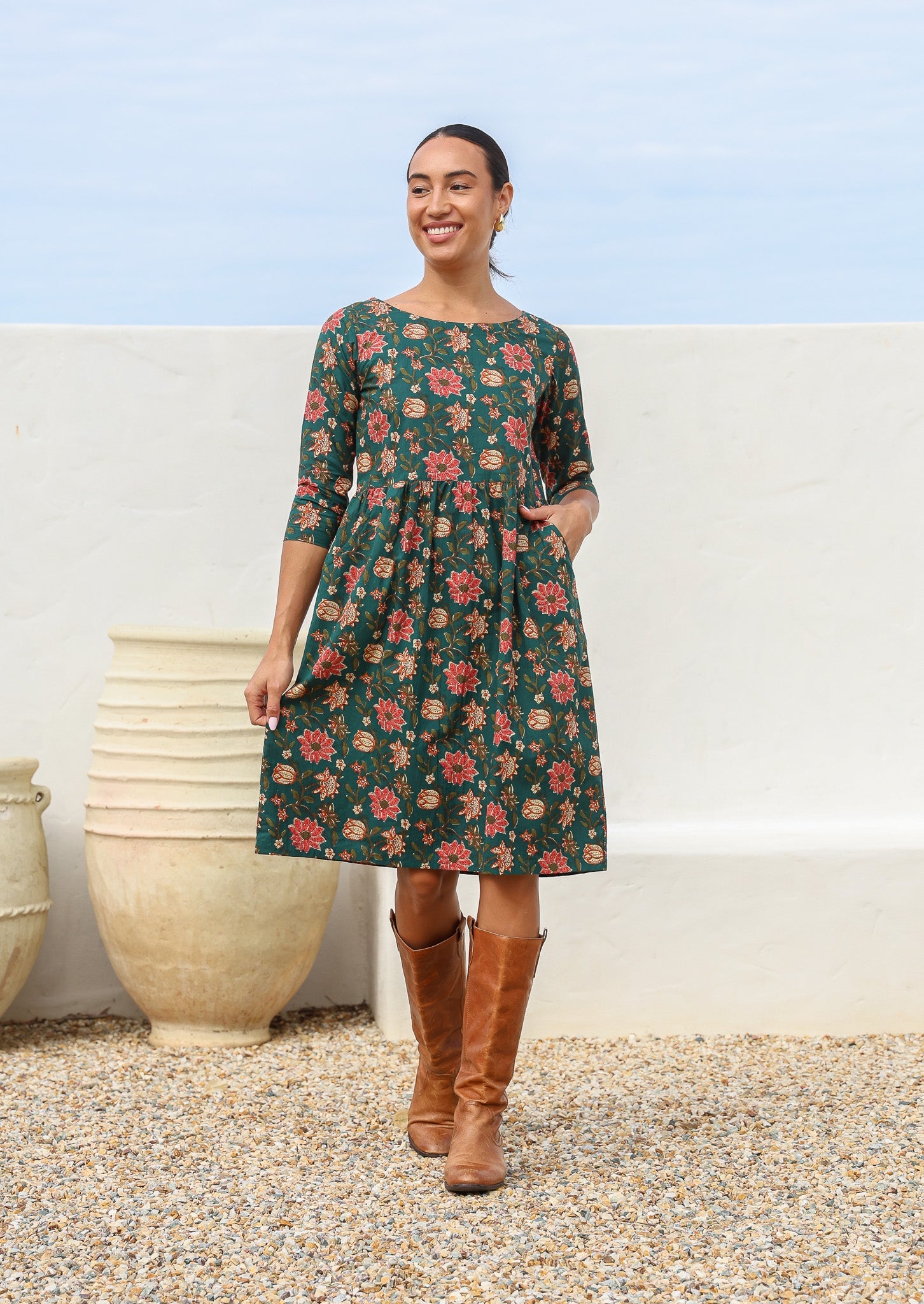 Woman wears 100% cotton knee length dress in deep green with beautiful pinkish red floral print and hand in pocket.