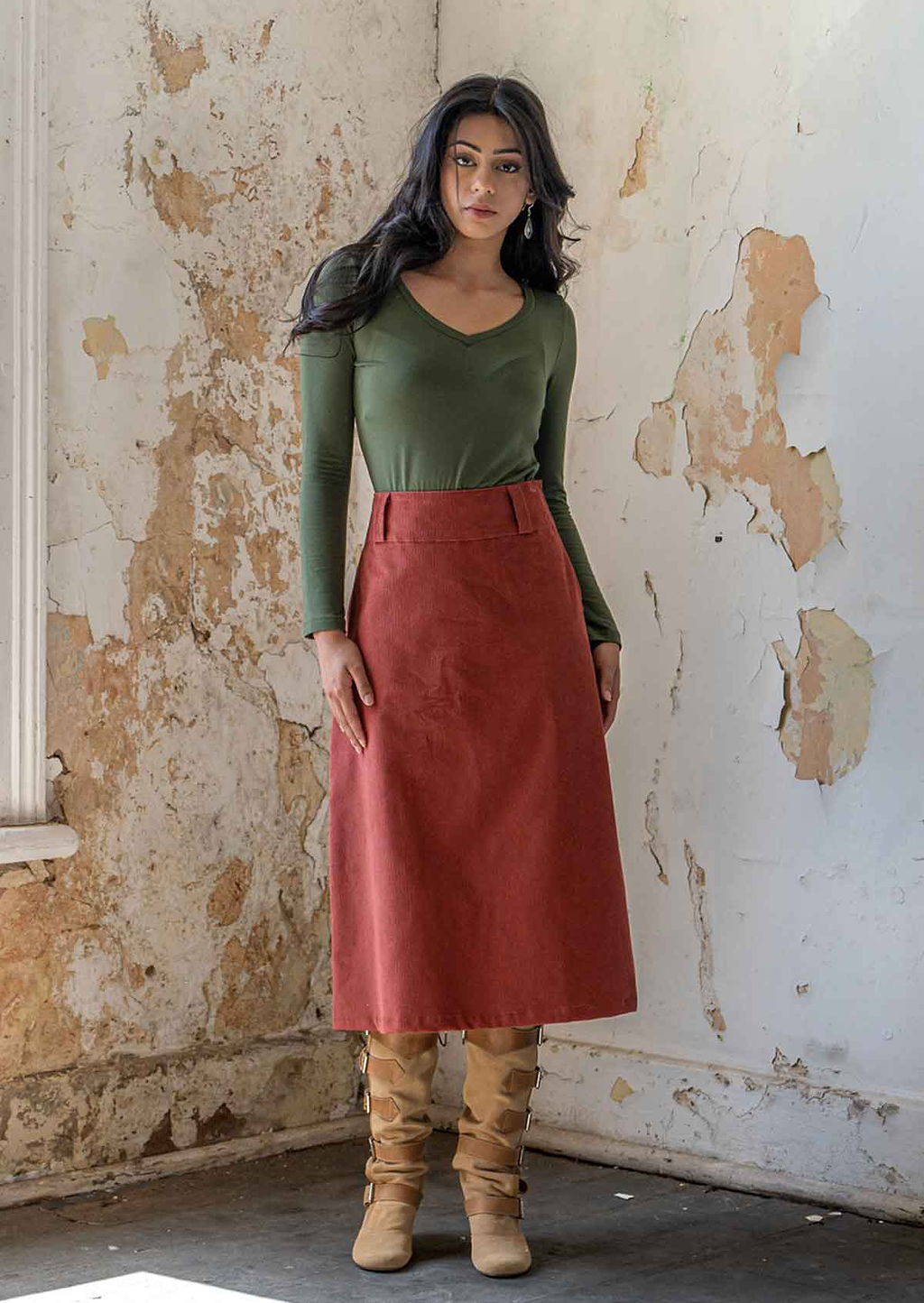 Woman wears burnt henna corduroy A-line midi length skirt with belt loops, paired with an olive green long sleeve top