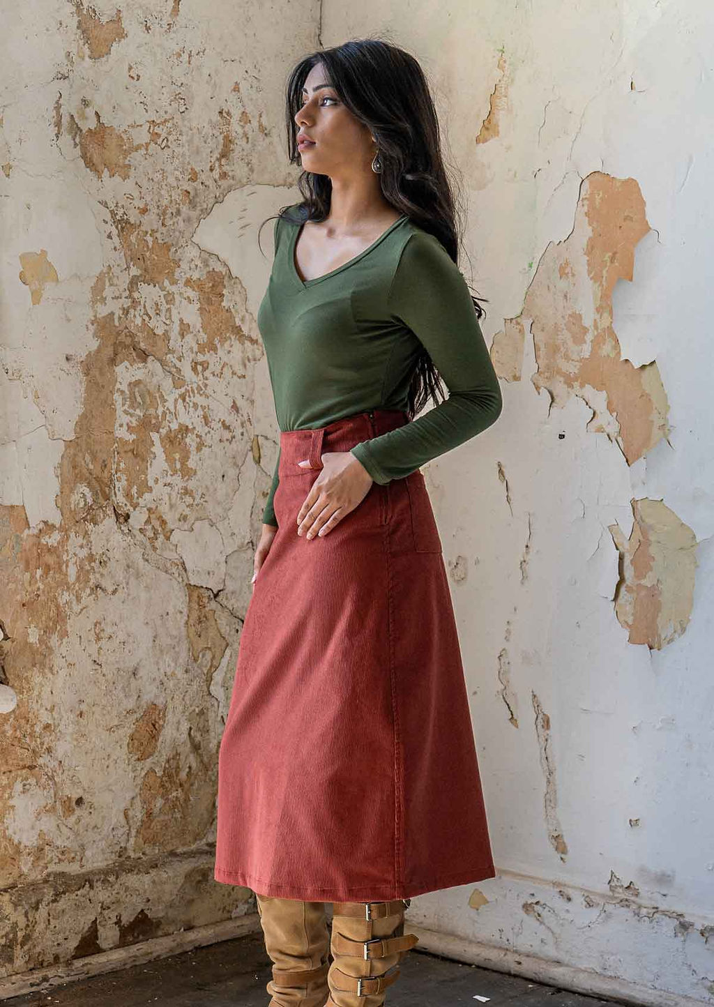 Woman stands side on in burnt henna coloured corduroy midi length skirt with thumb hooked in belt loop