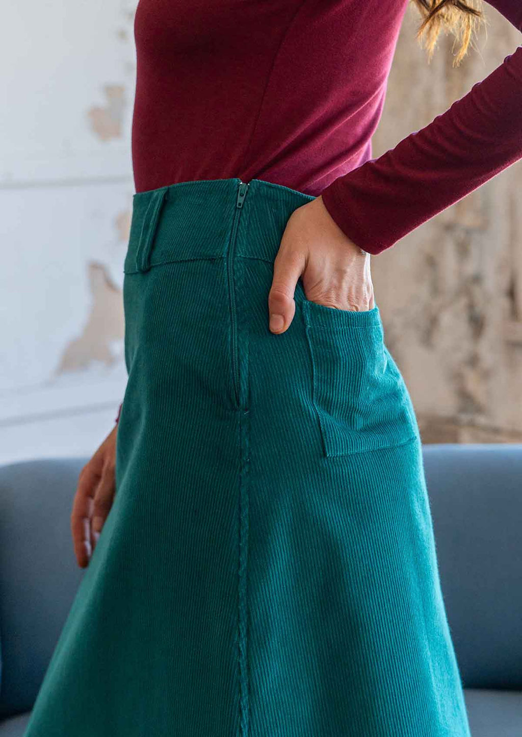 Close up of teal corduroy skirt with belt loops and side zip, with models hand in back pocket