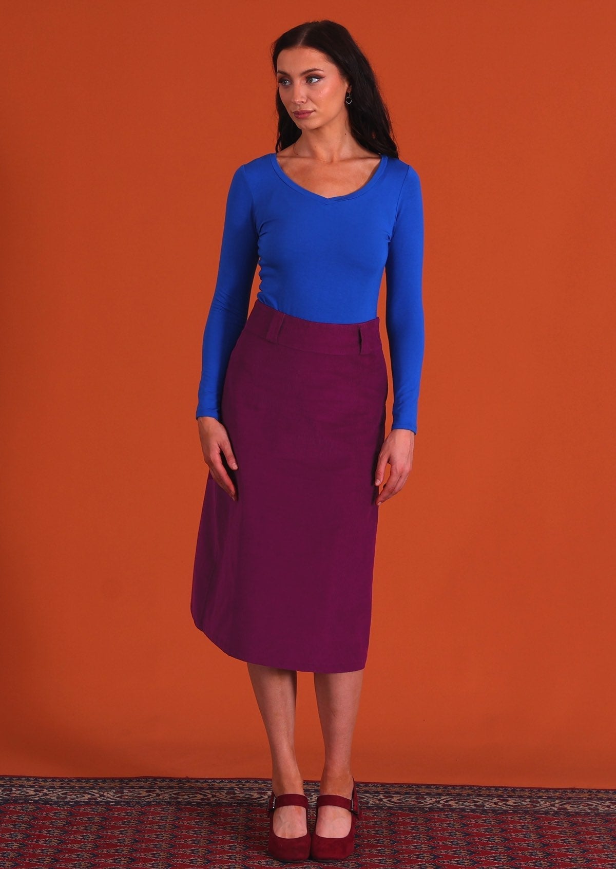 woman wearing purple corduroy aline skirt with belt loops with bright blue long sleeve top standing on persian carpet