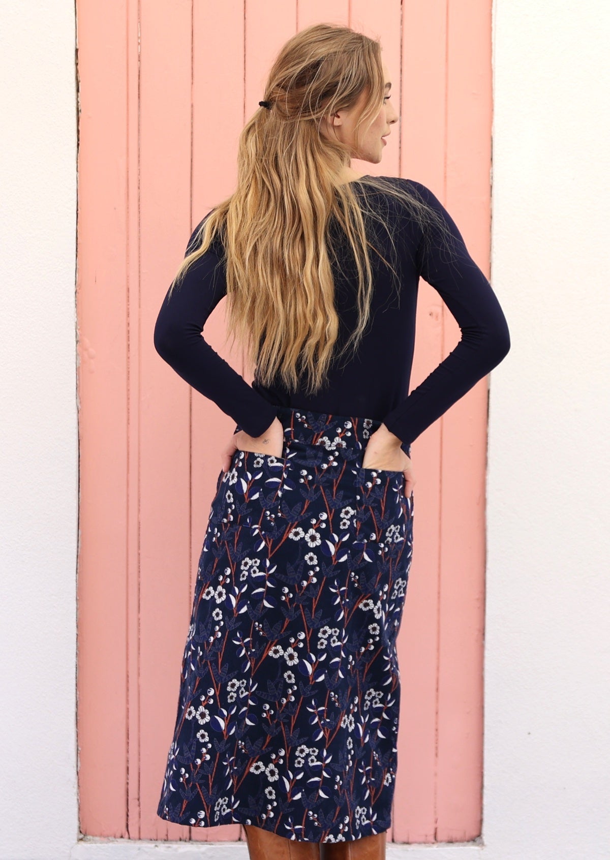Model wears a mid length floral printed corduroy skirt in navy blue and both the hands in back pockets
