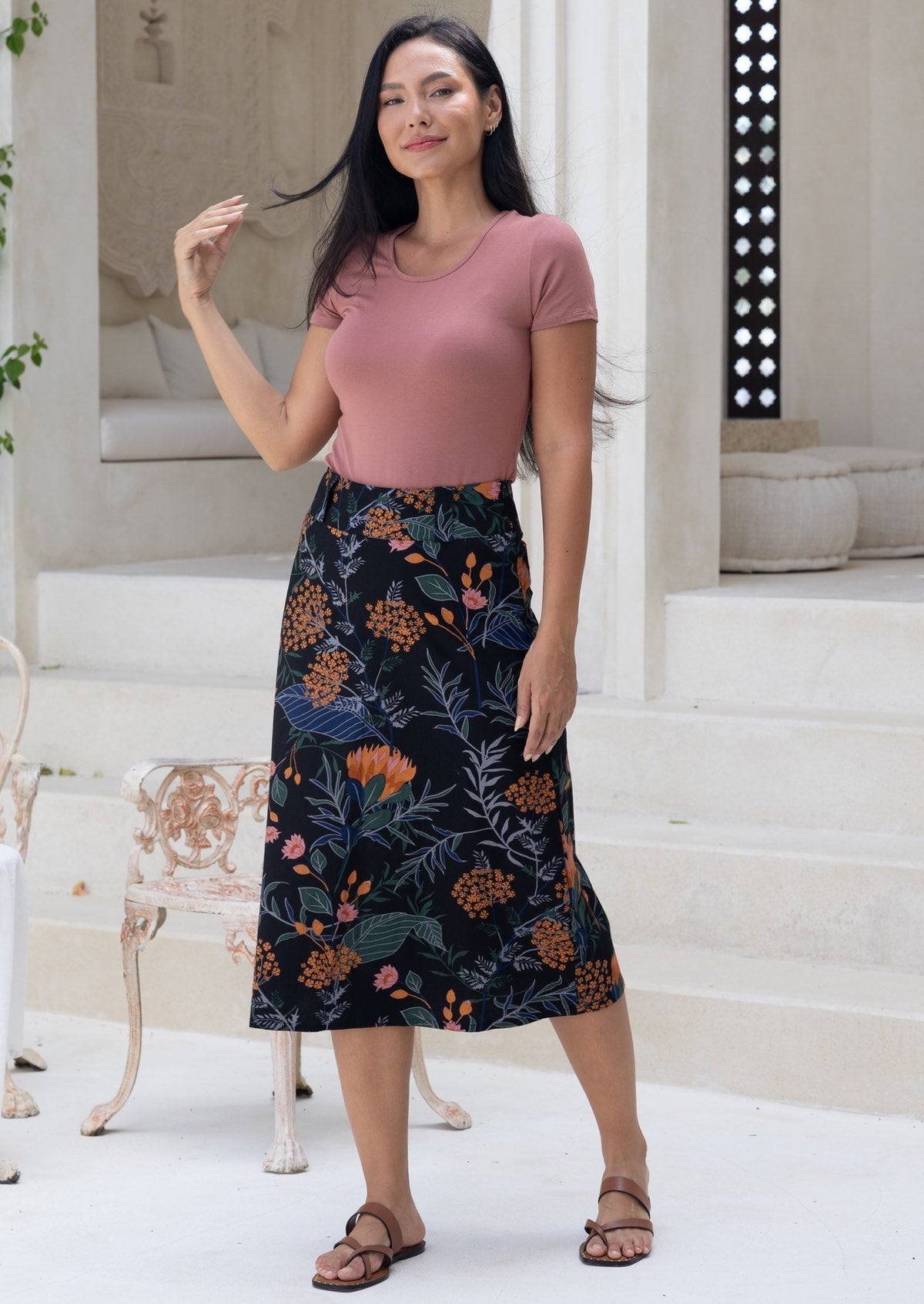 Woman wearing a pink top and a black based cotton floral shin length skirt standing in a light space