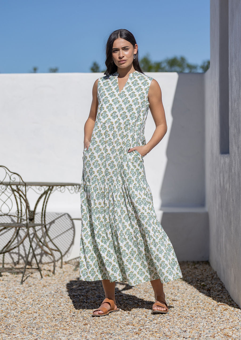 Woman wearing a sleeveless floral cotton maxi dress standing outdoors against a white wall.