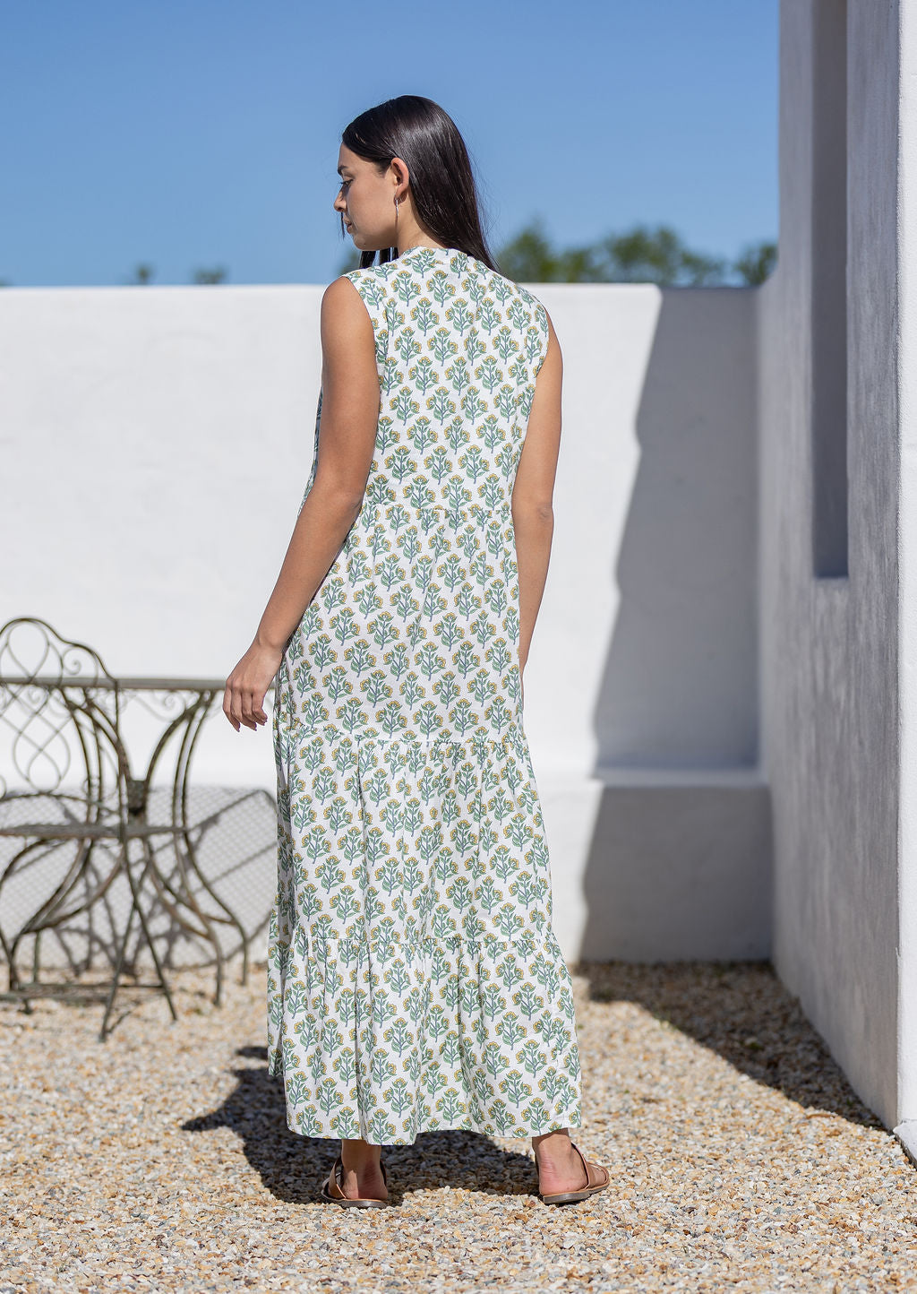 Woman wearing a white cotton patterned dress standing outdoors with a white wall and chair in the background.