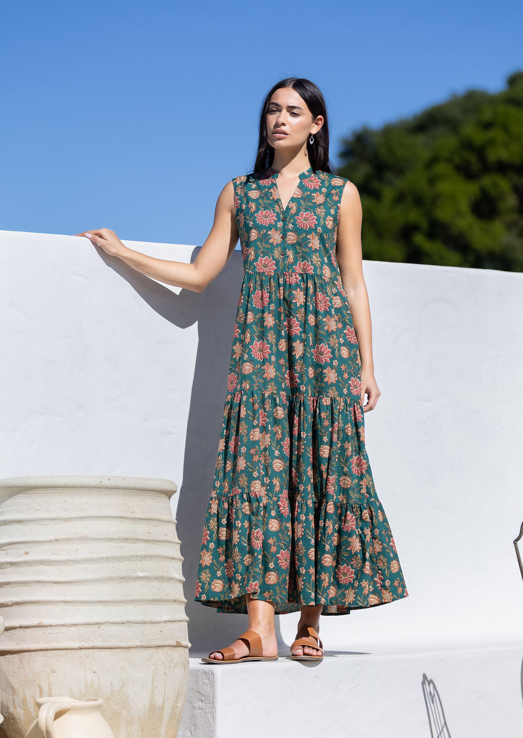 Woman wearing a green floral cotton maxi dress against a white wall with blue sky and greenery.