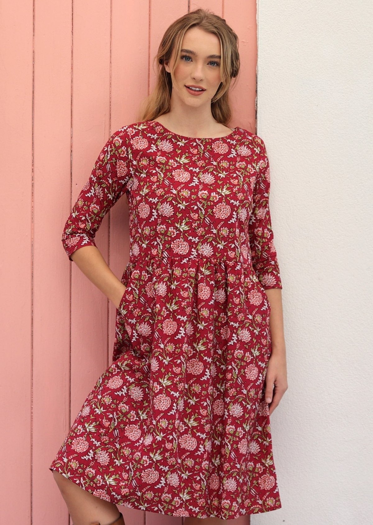 Model standing in front of pink wall wearing gorgeous cranberry based dress with simple round neckline.