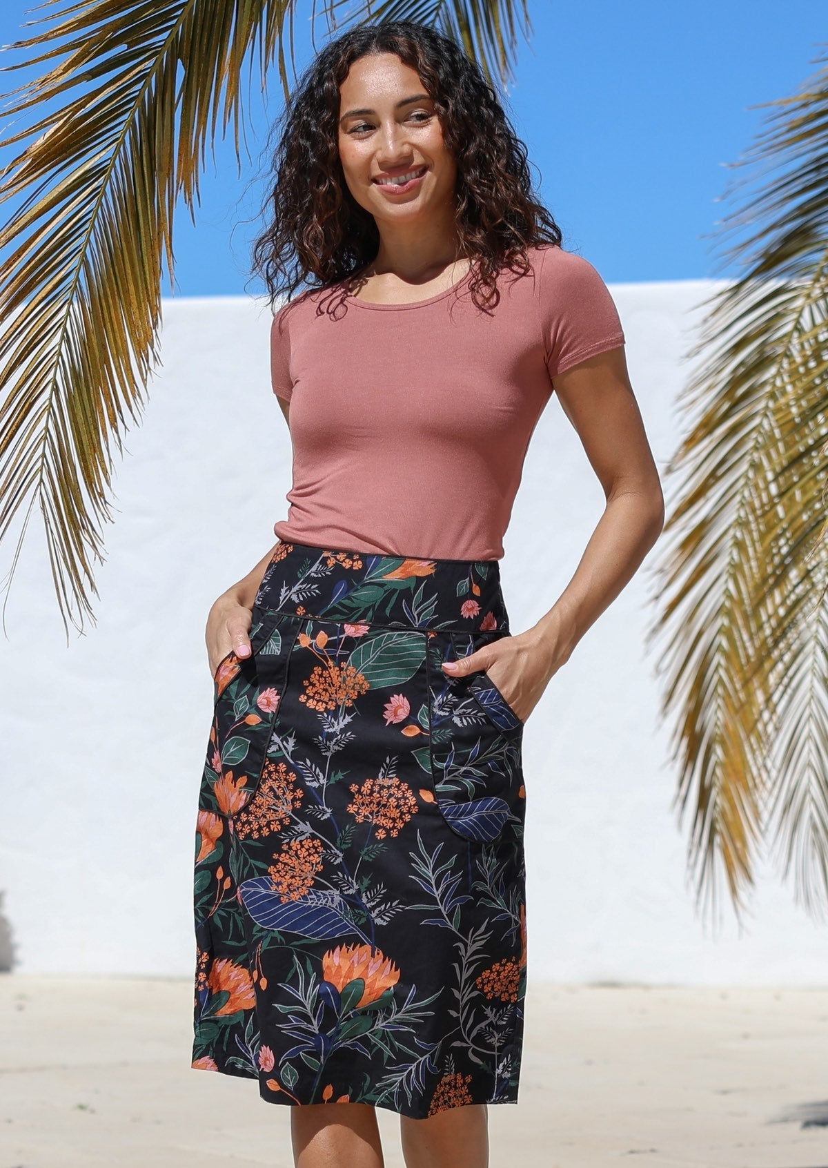 Woman wearing a high-waisted, knee length pencil skirt with piped detailing and pockets in a black based botanical print.