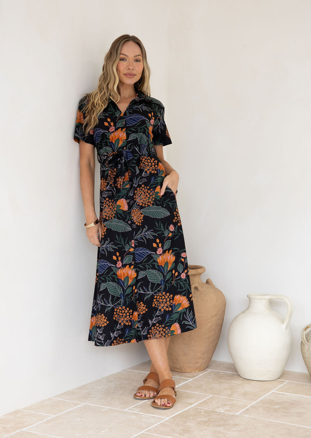 Model poses with one hand in pocket wearing a shirt style button through cotton dress with collar and a removable waist tie.