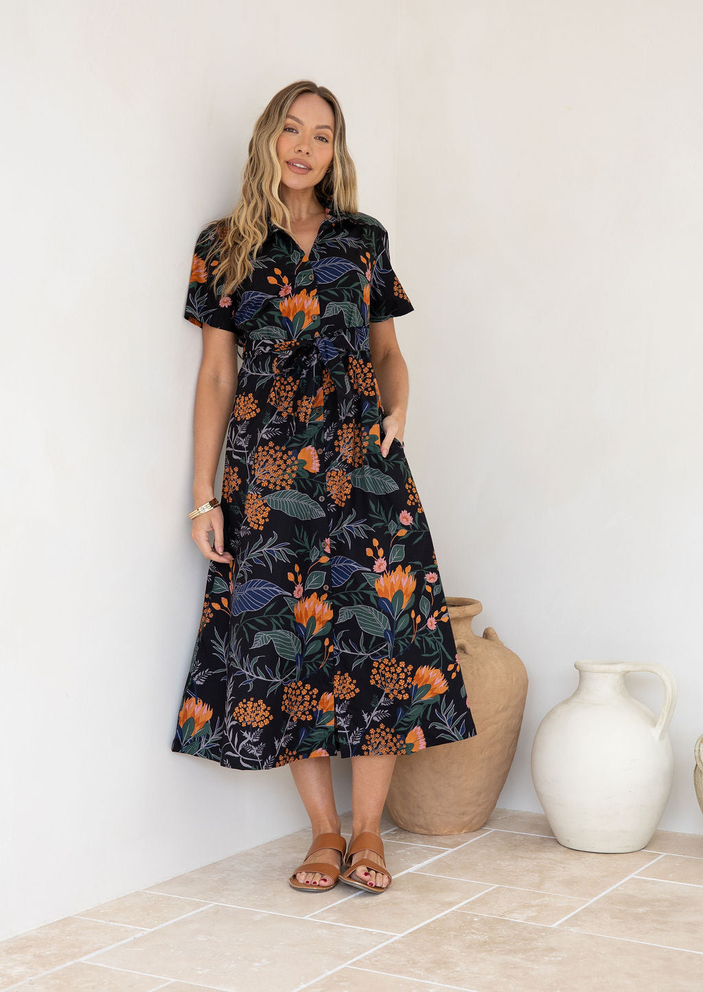 Woman wearing 100 percent cotton button through shirt dress with collar and short sleeves, in a moody botanical print on black base with pink and orange florals.