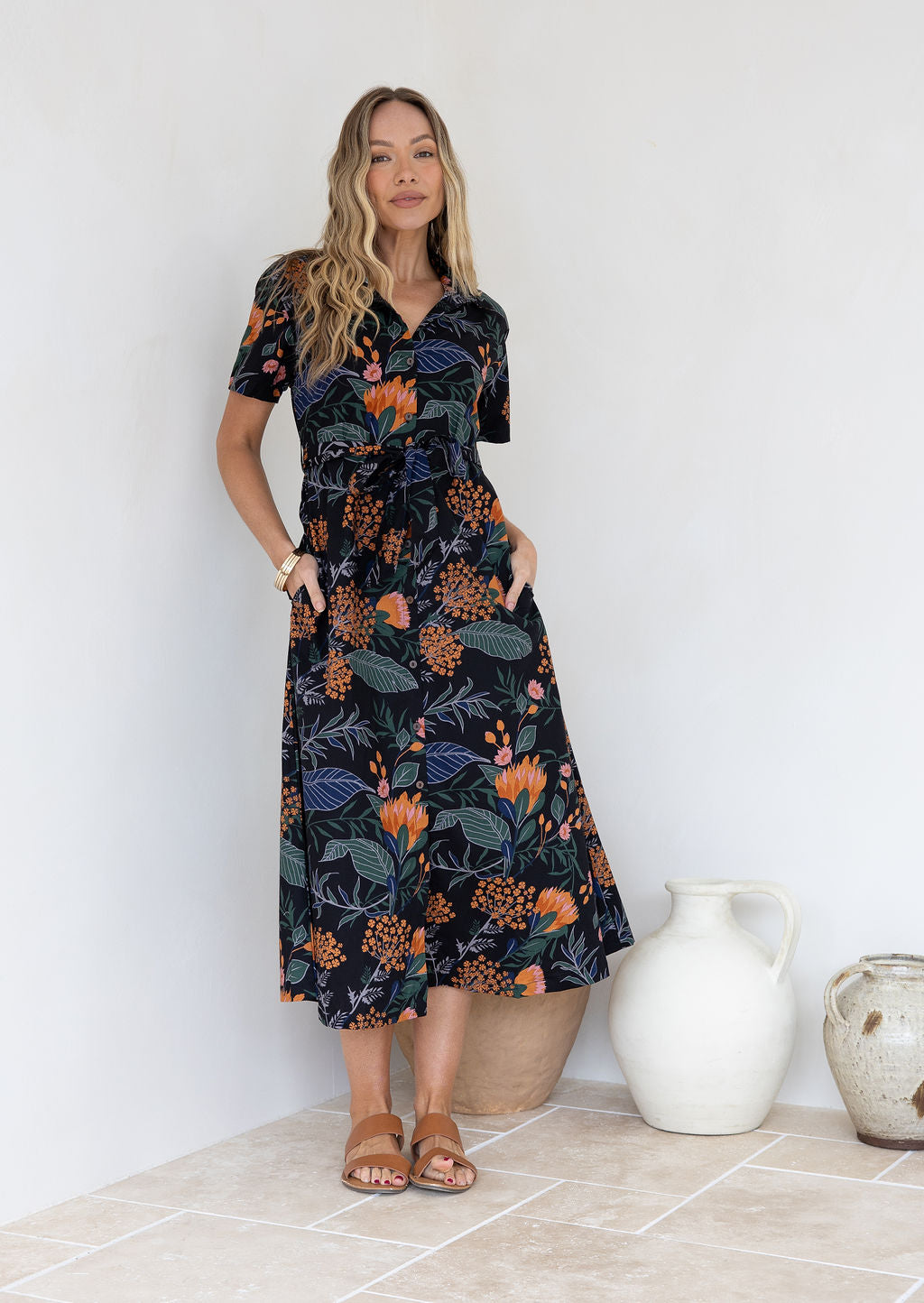 Model standing in front of white wall in floral printed shirt dress with detachable fabric belt, short sleeves and pockets.