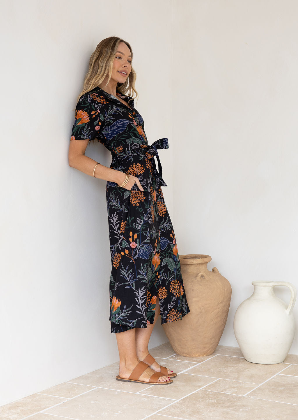 Model leaning on the wall wears 1950s inspired, button through shirt style dress with collar, short sleeves, waist tie and pockets.