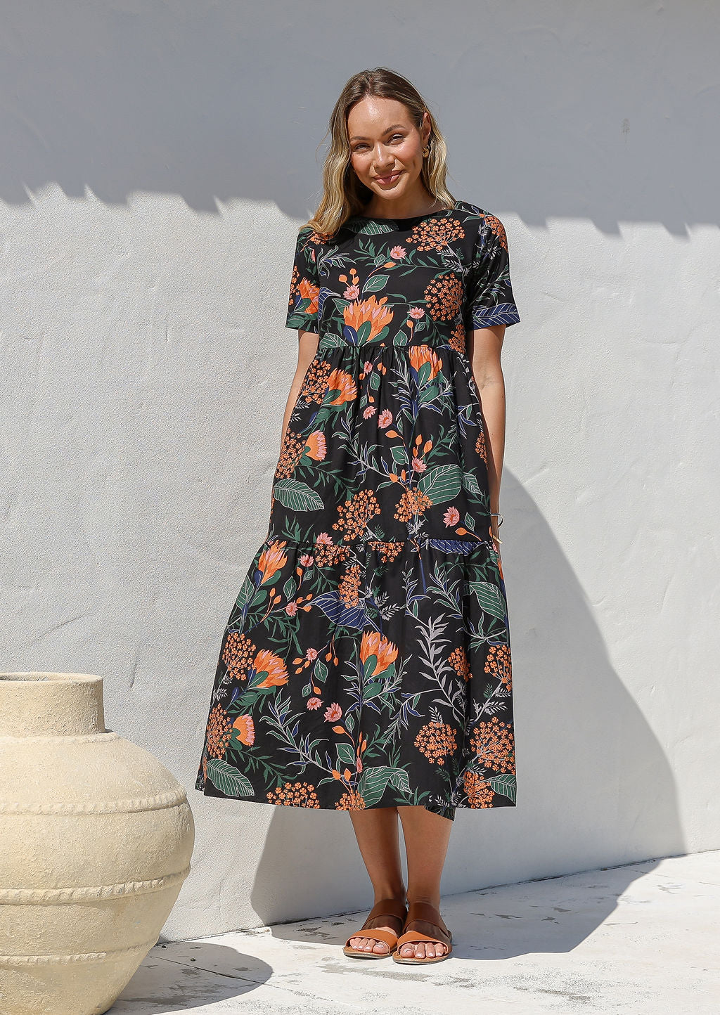 Woman wearing a 100% cotton, midlength black dress with botanical print featuring three tiered design, short sleeves, high round neckline and pockets
