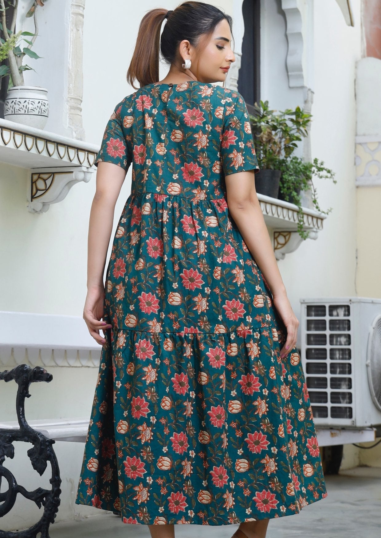 Back pose of model in 3 tiered, relaxed fit cotton dress with pockets and button back closure.