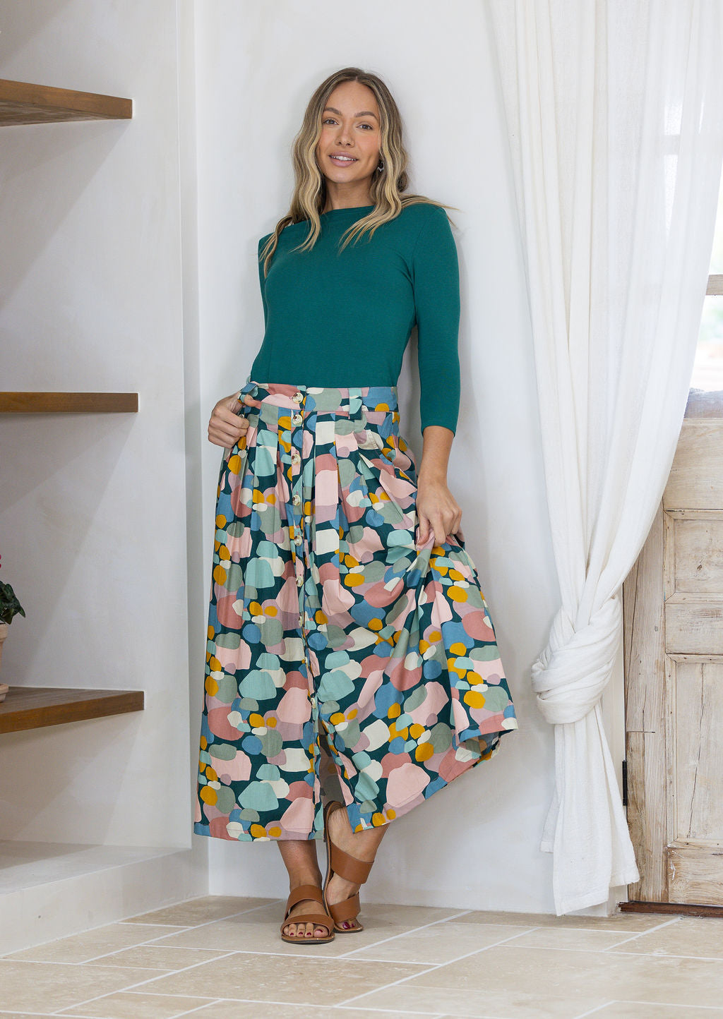 Woman wearing a green top and pastel spotty print button through skirt, leaning against a wall
