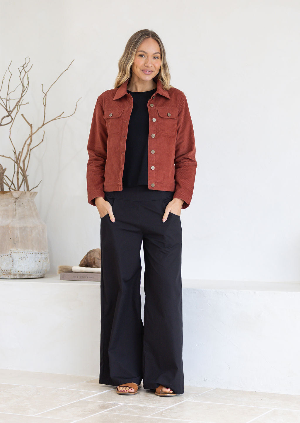 Woman wearing a red jacket and black cotton pants standing in a minimalistic room.