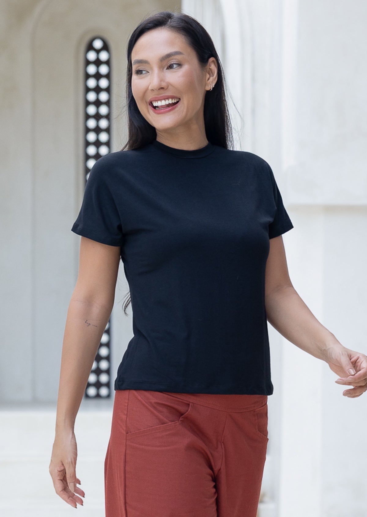 Woman wearing a black t-shirt and rust pants standing in front of a white wall with architectural elements.