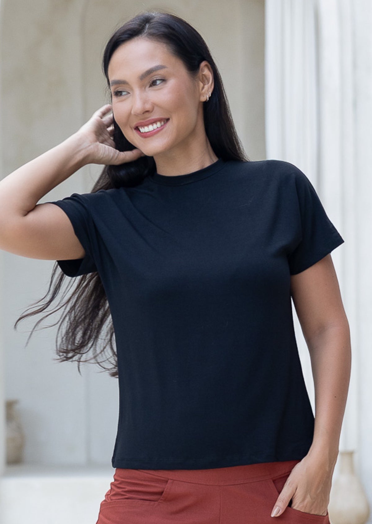 Woman wearing a black crewneck t-shirt and rust pants, swishing hair off shoulder
