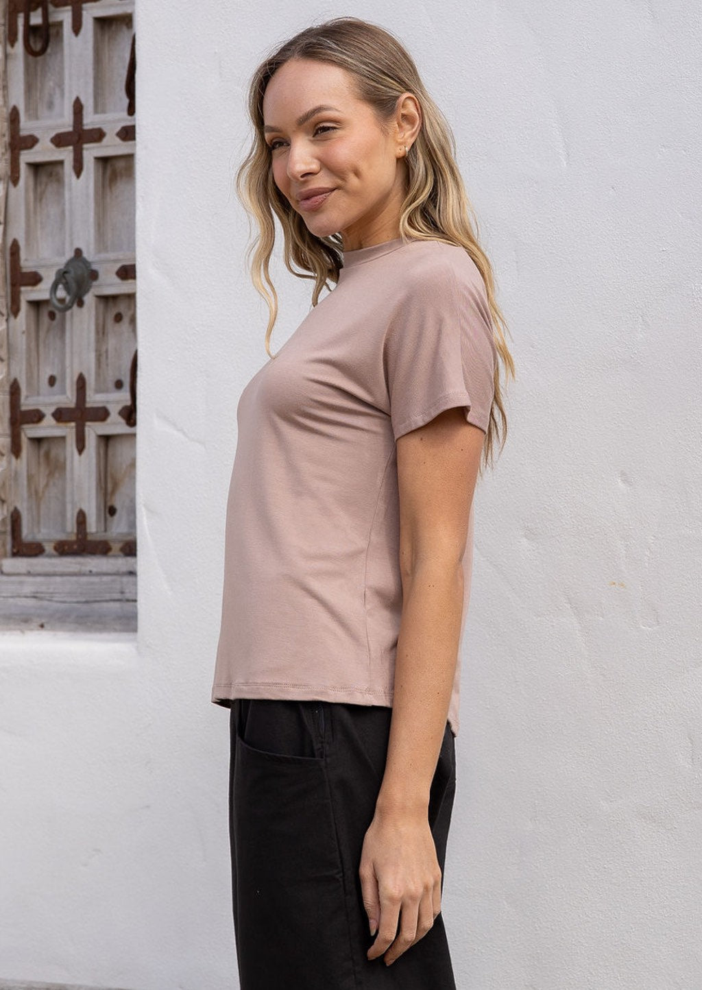 Woman standing side on wearing a beige t-shirt and black pants standing against a white wall.