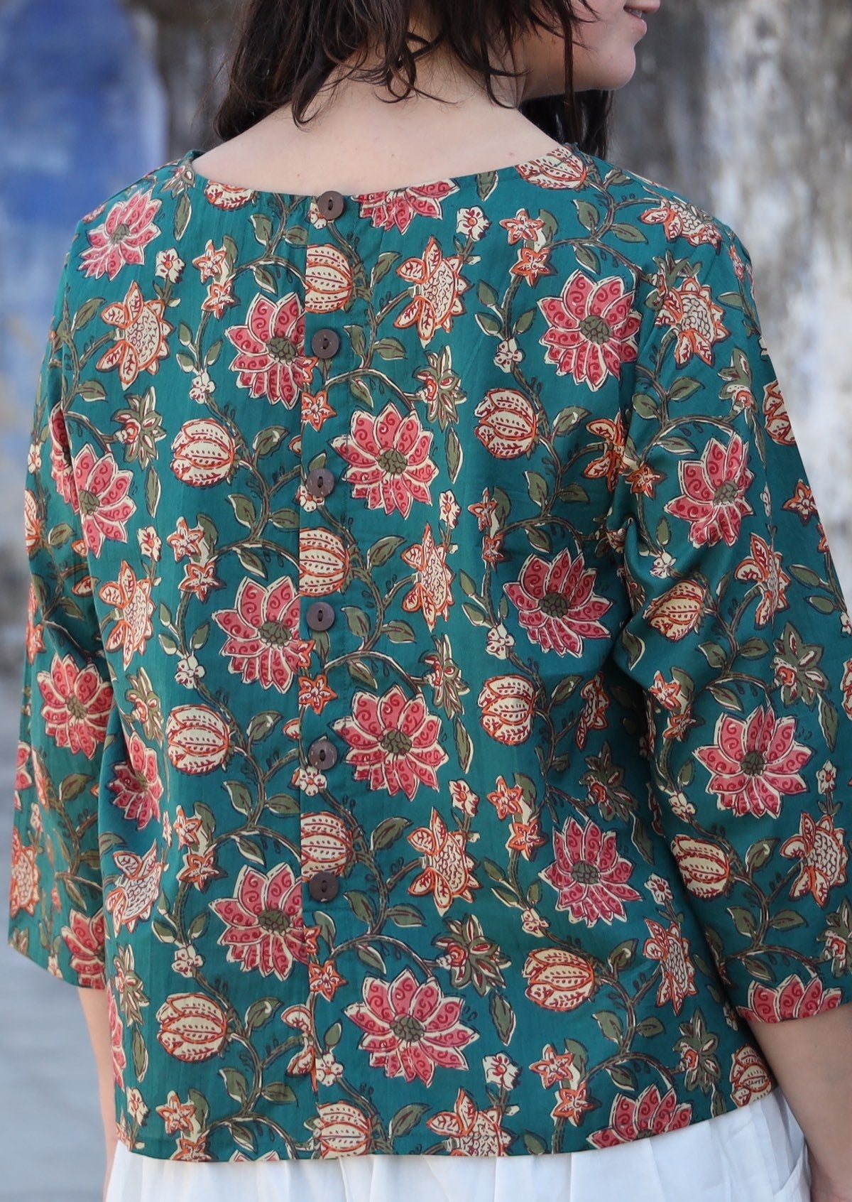 Back image of woman wearing relaxed fit cotton top with 3/4 sleeves and round neckline with decorative buttons on the back in green with a floral print