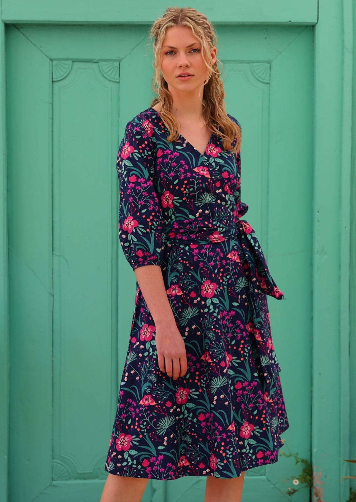 Blonde woman in long sleeve purple floral cotton wrap dress in front of aqua door