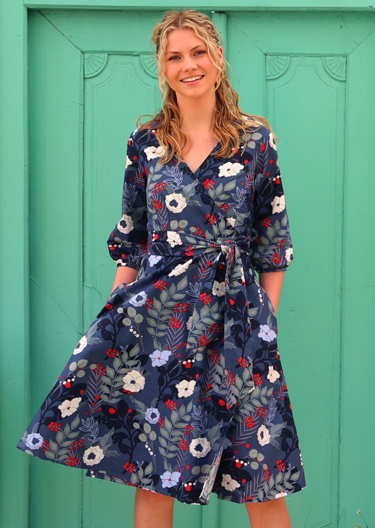 woman in 100% cotton wrap dress in blue with white flowers and long sleeves  with pockets in front of aqua door