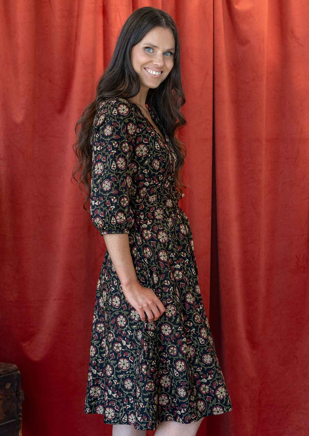 Model stands sideways wearing a black floral printed wrap dress with 3/4 sleeves, pockets and a waist-tie