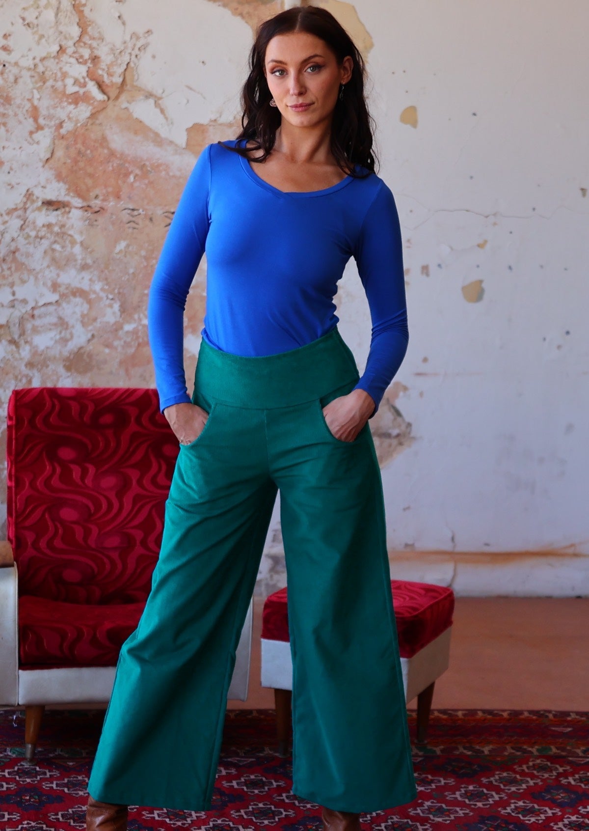 Woman wears electric blue long sleeve V-neck top with teal wide leg corduroy pants with a wide waistband, with her hands in pockets