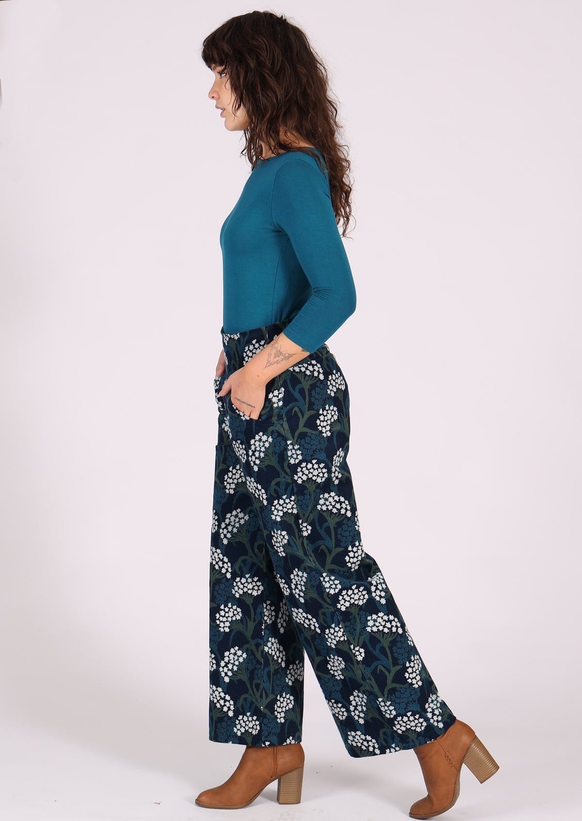 Woman stands side on in wide cord pants, paired with teal top and ankle boots, with hands in pockets