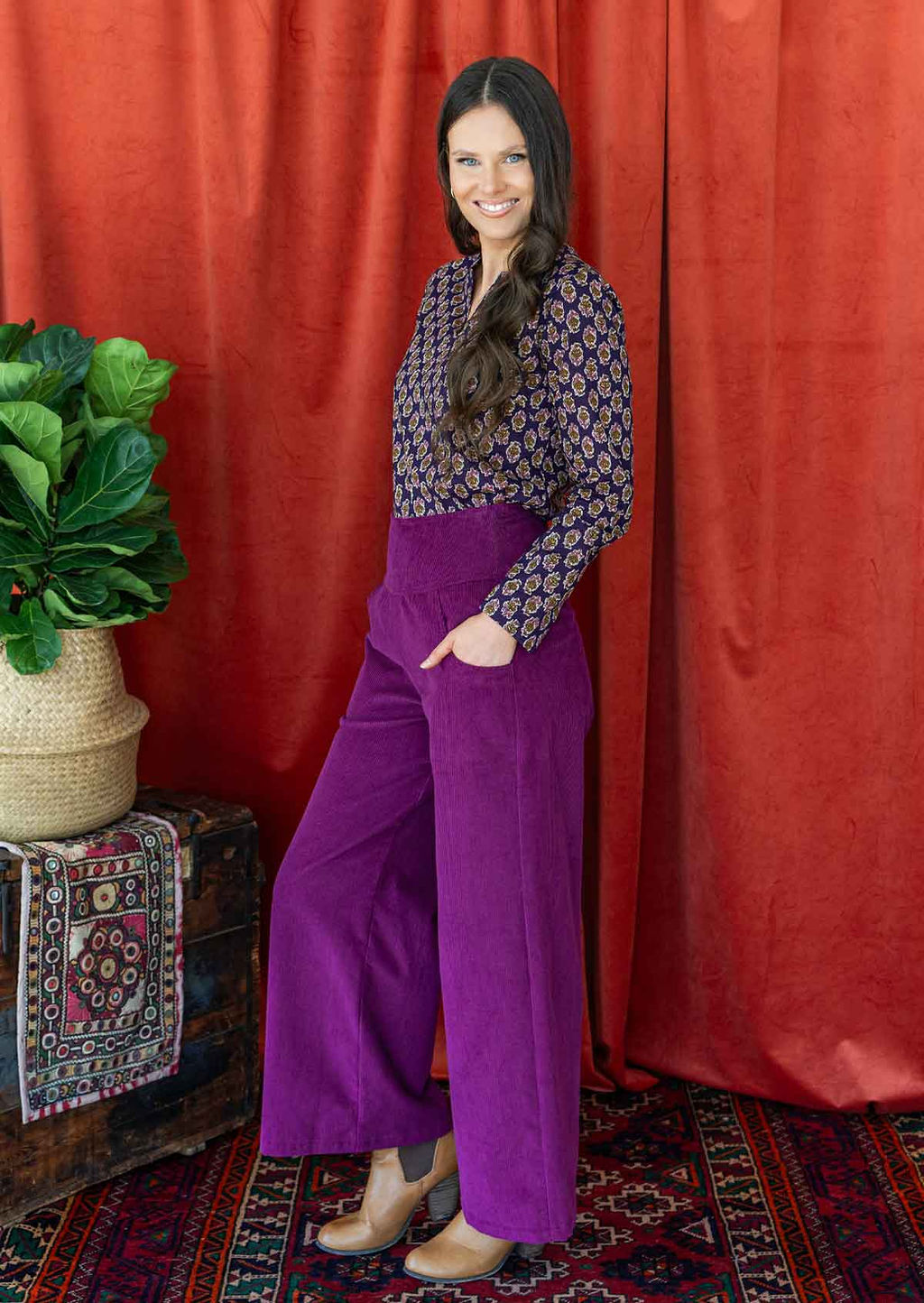 woman with long hair wears purple corduroy wide leg pants with her hand in pockets, paired with a long sleeve cotton blouse