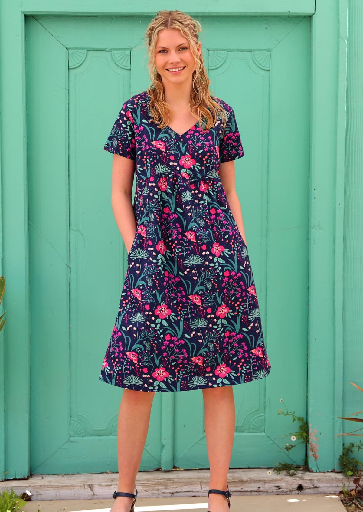 Model wearing 100 percent cotton v-neck sundress with hot pink and bright purple flowers on a deep blue base, with hands in pockets