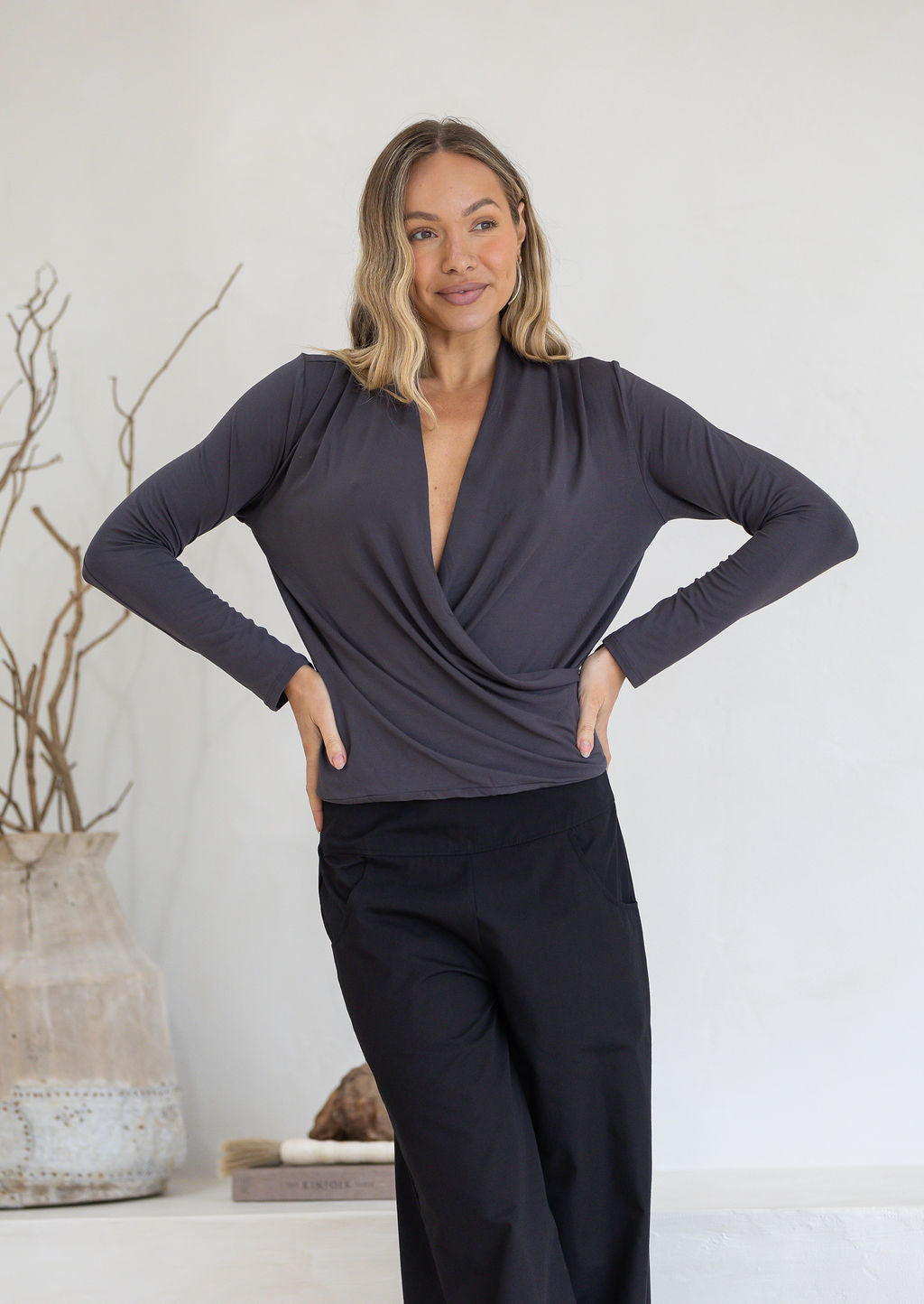 Woman wearing a dark grey long sleeve crossover top with pleats at the front of the shoulders with black pants