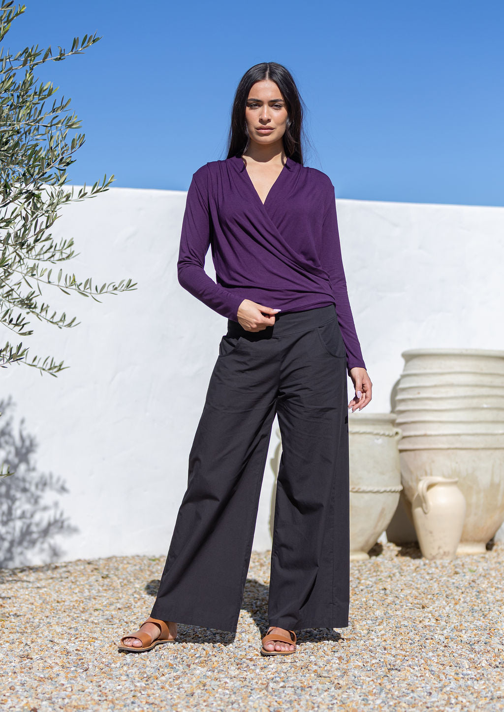 Woman wearing a purple crossover long sleeve top and black pants standing in front of a white wall