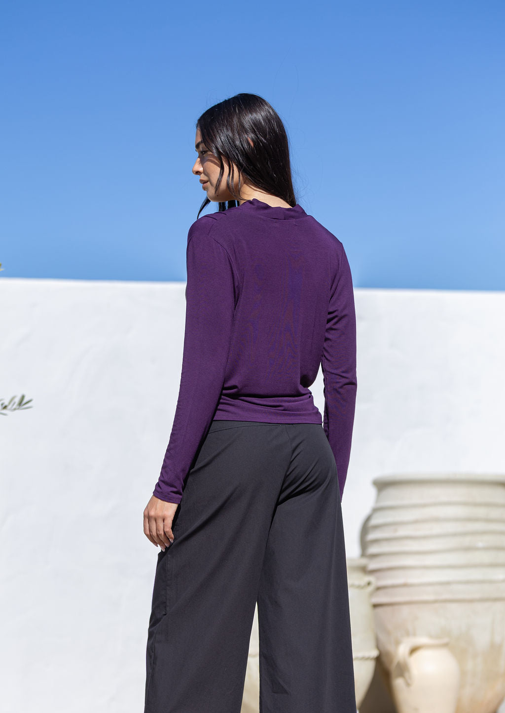 Back view of woman wearing a purple long sleeve top and dark pants against a blue sky.
