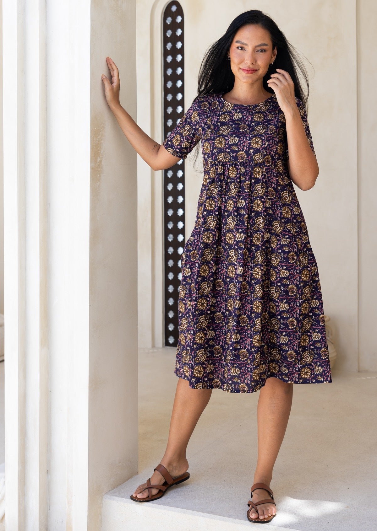 Woman wearing a purple floral sun dress standing against a white wall.